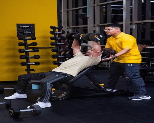 Portrait of Luis G., gym floor trainer assisting a beginner with weight machines
